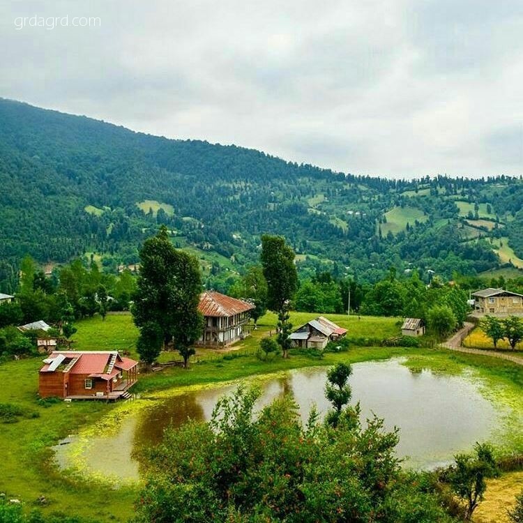 منظره‌ای زیبا از یک روستای سرسبز در شمال ایران با خانه‌های سنتی و یک برکه‌ی آبی در پیش زمینه، در میان کوه‌های سرسبز