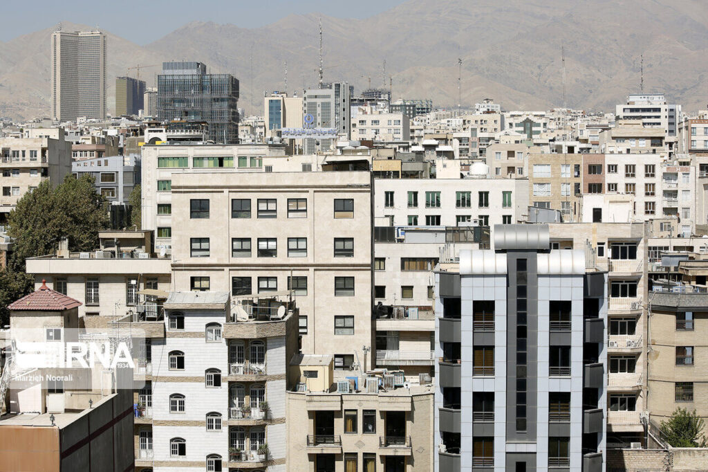 تصویری از یک منظره شهری با ساختمان‌های مختلف و متنوع در مناطق مختلف. در پس‌زمینه، کوه‌ها و آسمان صاف دیده می‌شوند