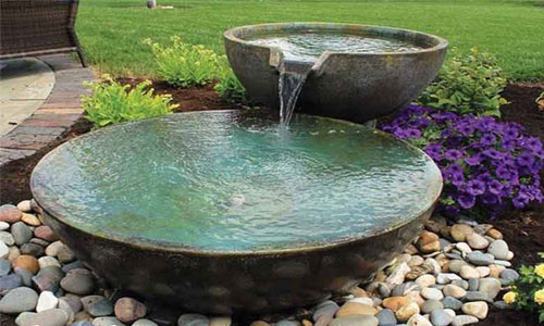 یک آبنمای مدرن دو کاسهای با جریان آب از کاسه بالایی به پایینی، احاطه شده با سنگهای رودخانهای رنگارنگ