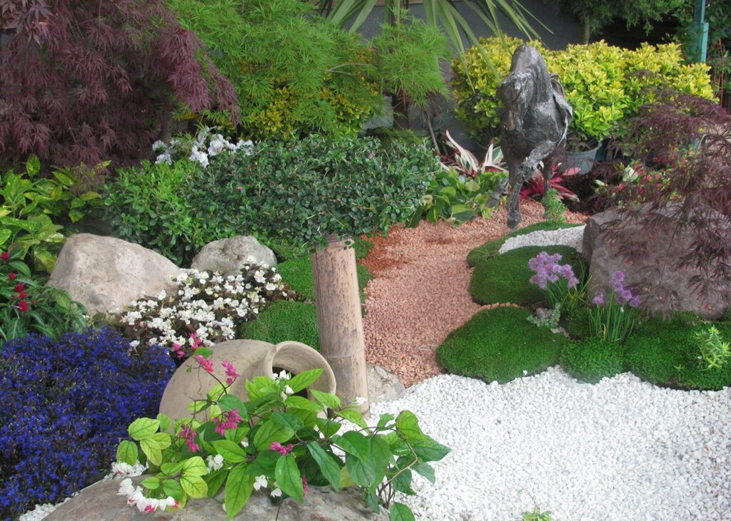 یک باغ صخرهای کوچک و پر از گیاهان متنوع با رنگها و بافتهای مختلف، سنگهای بزرگ، یک کوزه سفالی شکسته و یک مجسمه فلزی تیره