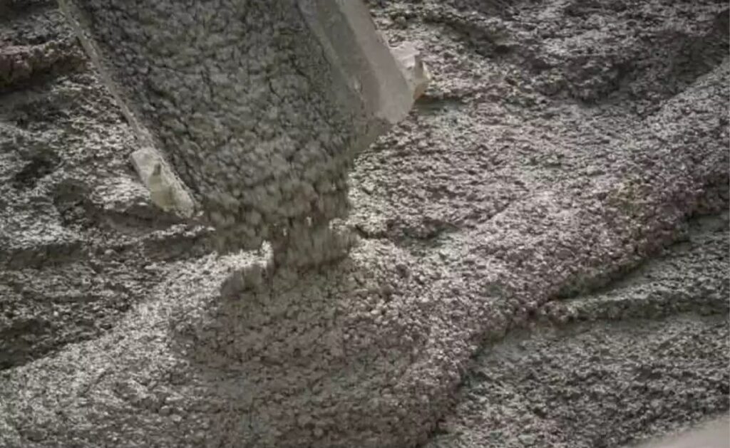 بتن تازه یا ملات سیمانی خاکستری رنگ در حال ریخته شدن از یک ناودان یا خروجی به روی تودهای از همین ماده، با بافتی دانهدار و مرطوب