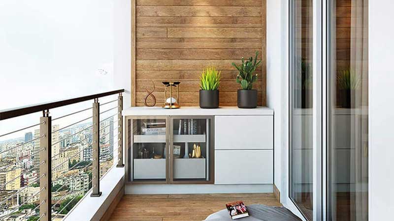 بالکن کوچک و مدرن با کفپوش چوبی، یک کابینت سفید با درهای شیشهای و قفسههای چوبی، دو گلدان مشکی با گیاهان سبز مصنوعی، یک شمعدان طلایی مدرن و یک کوسن خاکستری روی زمین