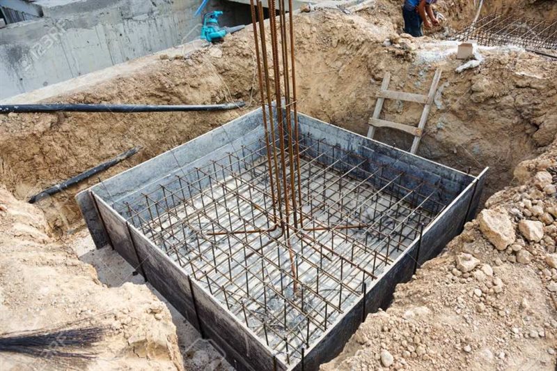 قالب مربعشکل فونداسیون بتنی با شبکهای از میلگردهای فولادی و میلگردهای عمودی بیرون زده در یک گودال خاکی در یک سایت ساختمانی