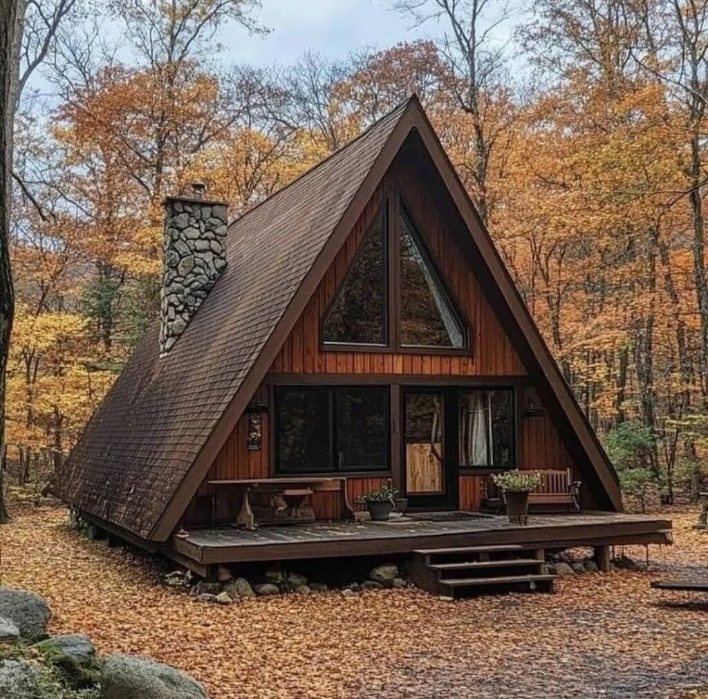 کلبه A-Frame چوبی سنتی در جنگلی پاییزی با سقف قهوهای تیره و دودکش سنگی