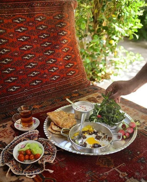 میز صبحانه سنتی ایرانی روی فرشی قرمز با تخممرغ نیمرو، نان، سبزی تازه، تربچه، ماست و خیار و گوجه در فضای باز