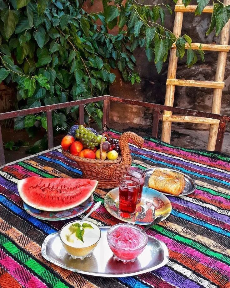 میز صبحانه یا عصرانه سنتی ایرانی در فضای باز با فرش دستباف رنگارنگ، میوههای تازه، هندوانه، نوشیدنی و دو کاسه دسر 1