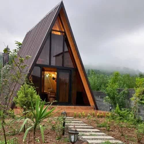 یک کلبه مدرن سوئیسی (A-frame) با نمای چوبی و پنجرههای بزرگ شیشهای در میان منظرهای سرسبز و کوهستانی با مه