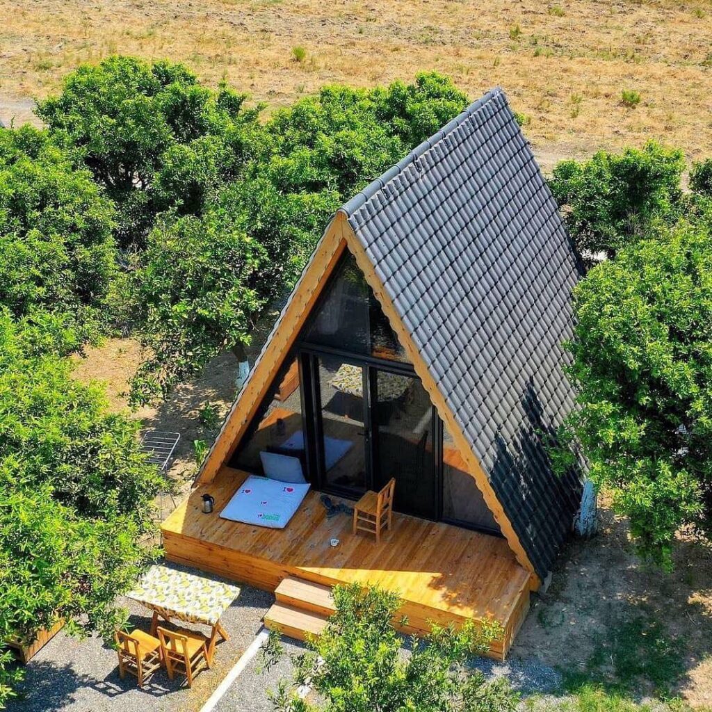 یک کلبه مدرن سوئیسی (A-frame) با سقف کاشیکاری شده خاکستری و نمای چوبی، در میان باغی سرسبز با درختان میوه