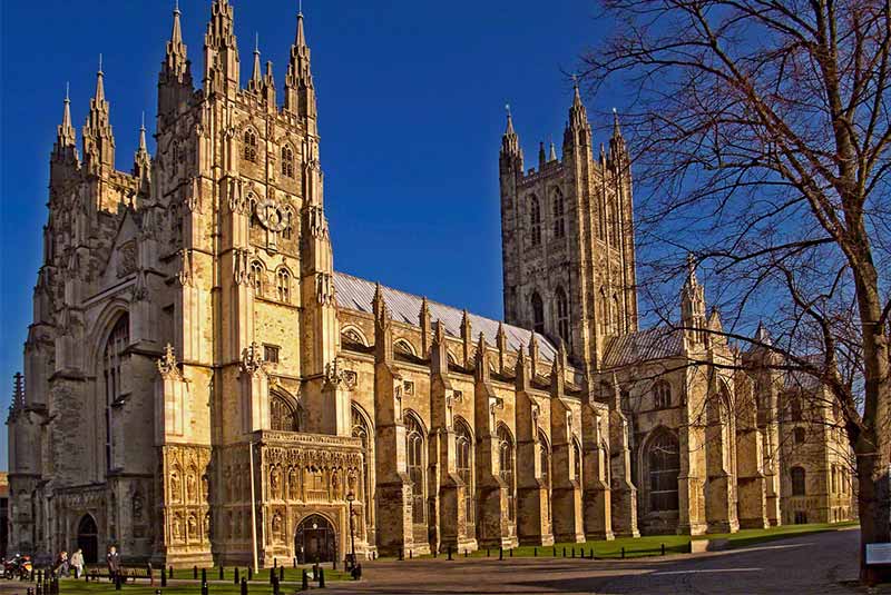 تصویری خارجی از کلیسای جامع کانتربری، سازهای عظیم و باشکوه به سبک معماری گوتیک، با برجهای بلند، پنجرههای کشیده و تزئینات سنگی ظریف، تحت تابش نور خورشید و آسمانی آبی