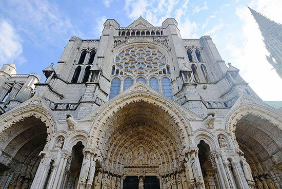 تصویری از نمای غربی کلیسای جامع شارتر (Chartres Cathedral) با تمرکز بر ورودیهای اصلی و پنجره گرد (روزتا) بزرگ بالای آن. نمای سنگی با مجسمههای فراوان، طاقهای نوکتیز و یک برج بلند نامتقارن، نمونهای شاخص از معماری گوتیک اولیه و عالی فرانسه است