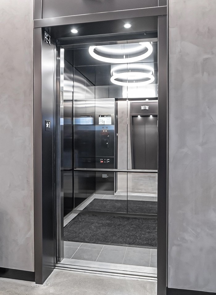 درب آسانسور باز شده در یک لابی با دیوارپوش خاکستری بتنی. داخل کابین آسانسور دارای دیوارهایی از جنس فولاد ضدزنگ بسیار براق (آینه‌ای) به رنگ دودی یا مشکی است