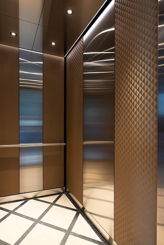 کابین آسانسور لوکس با طراحی ترکیبی از فولاد ضدزنگ براق (آینه‌ای) و پنل‌های فلزی یا کامپوزیتی با روکش برنزی/قهوه‌ای مات و طرح‌دار