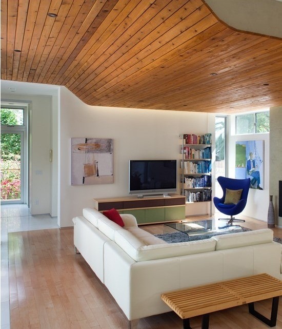 یک اتاق نشیمن مدرن با سقف کاذب چوبی منحنی و تخته‌ای، دیوارهای سفید و کف چوبی روشن. مبلمان شامل یک مبل سکشنال سفید، میز عسلی شیشه‌ای و یک قفسه کتاب فلزی است