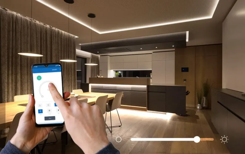 تصویری از کنترل نورپردازی هوشمند یک آشپزخانه و ناهارخوری مدرن (Open Plan) از طریق اپلیکیشن موبایل، با سقف کاذب لایه‌ای و نورپردازی مخفی LED و آویزهای مدرن