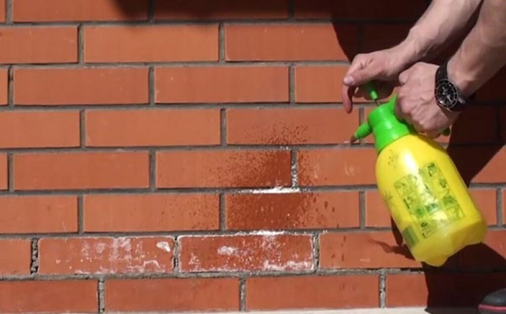 پاشش محلول پاک‌کننده (احتمالاً محلول سرکه یا اسید رقیق) بر روی نمای آجری دارای شوره سفید (Efflorescence) برای از بین بردن رسوبات نمک