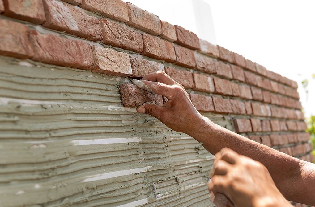 نصب دستی یک تایل آجری نازک با بافت متخلخل توسط کارگر روی لایه‌ای ضخیم از چسب ملات دندانه‌دار که برای اجرای نمای آجری سبک آماده شده است